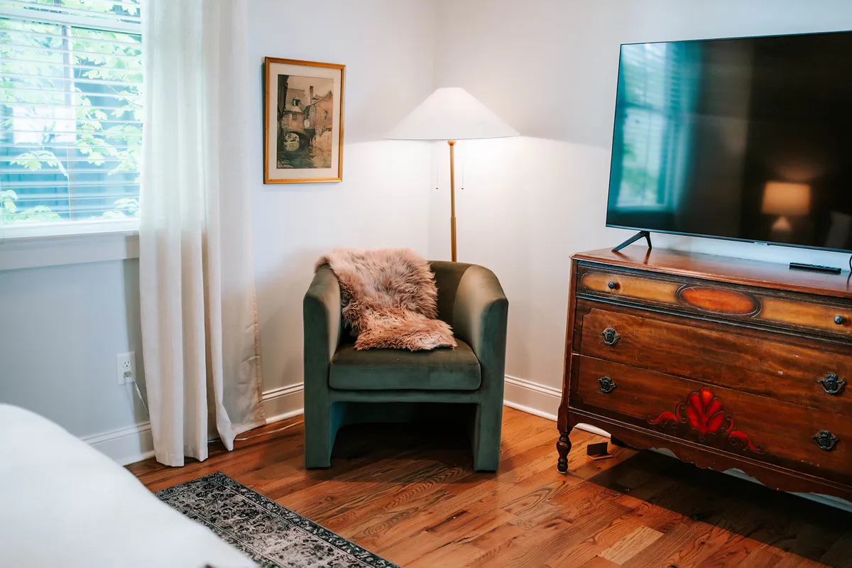 A welcoming living area featuring a green armchair beside a wooden dresser, a stylish lamp, and framed artwork. The space appears bright and inviting, perfect for relaxing, reading, or spending quiet moments together.