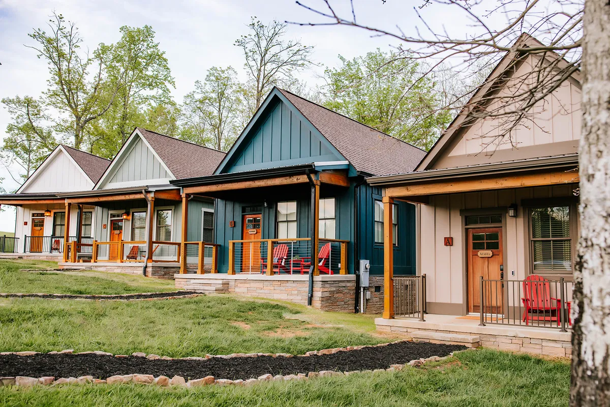 A view of colorful cottages lining a landscaped pathway, with bright red rocking chairs on the porches. The setting invites relaxation, nature walks, and casual gatherings with neighbors, framed by lush greenery and sunset lighting.
