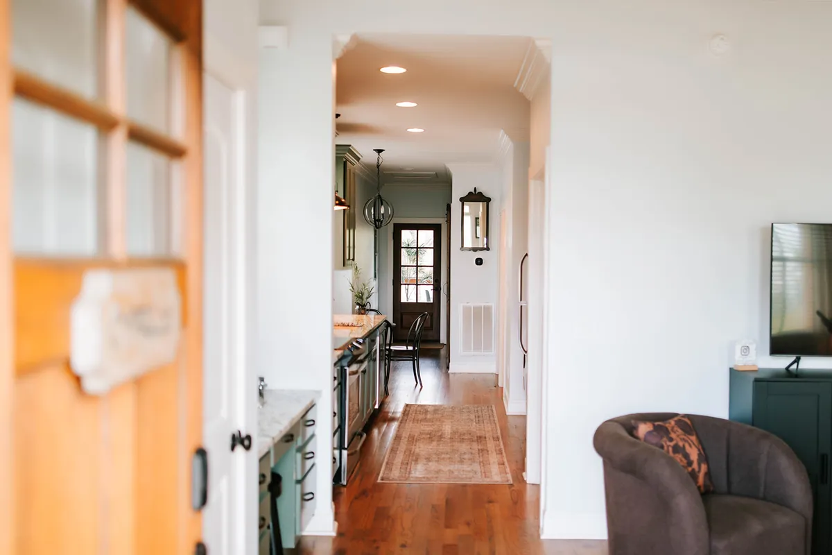 This wide interior shot encompasses the open-plan living area, showing a stylish living room with comfortable seating, a large wooden front door, a modern kitchen island, and a connected dining area. Sunlight streams in through expansive windows, illuminating the inviting communal space. The design incorporates warm wood tones and tasteful decor, embodying a blend of Prairie School Craft style and contemporary comfort.