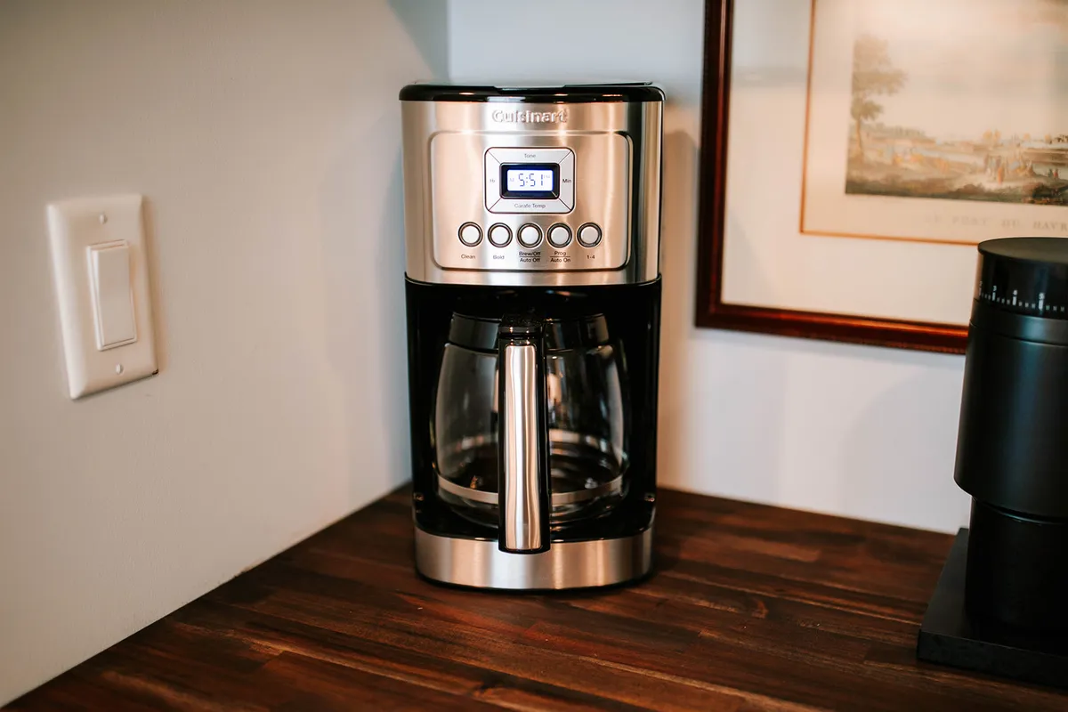 A kitchen scene with a coffee maker on a wooden countertop, accompanied by warm-toned framed artwork and a black cylindrical object. The ambiance is welcoming, suggesting morning rituals or catching up with friends before heading out for activities.