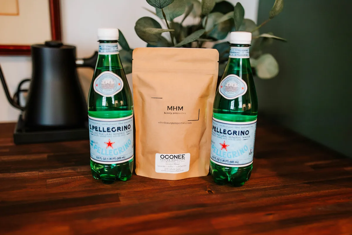 A close-up of two bottles of sparkling water and a brown paper bag labeled 'MHM,' set against a background with greenery. The scene suggests refreshment and casual gatherings—moments enjoyed before or after venturing out.