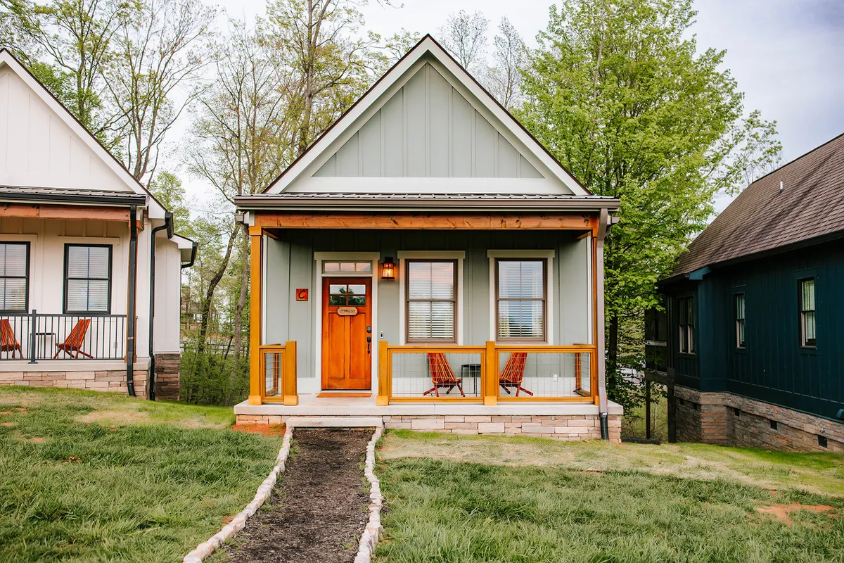 The hero image showcases the exterior of a chic, single-level cabin with inviting green siding, a charming wooden door, and a spacious front porch. A rock path leads to the entrance, and the cozy porch offers a perfect spot for sipping coffee at sunrise, unwinding at sunset, or gathering with friends. The scene feels both welcoming and tranquil, set against the natural beauty of Jasper Highlands.