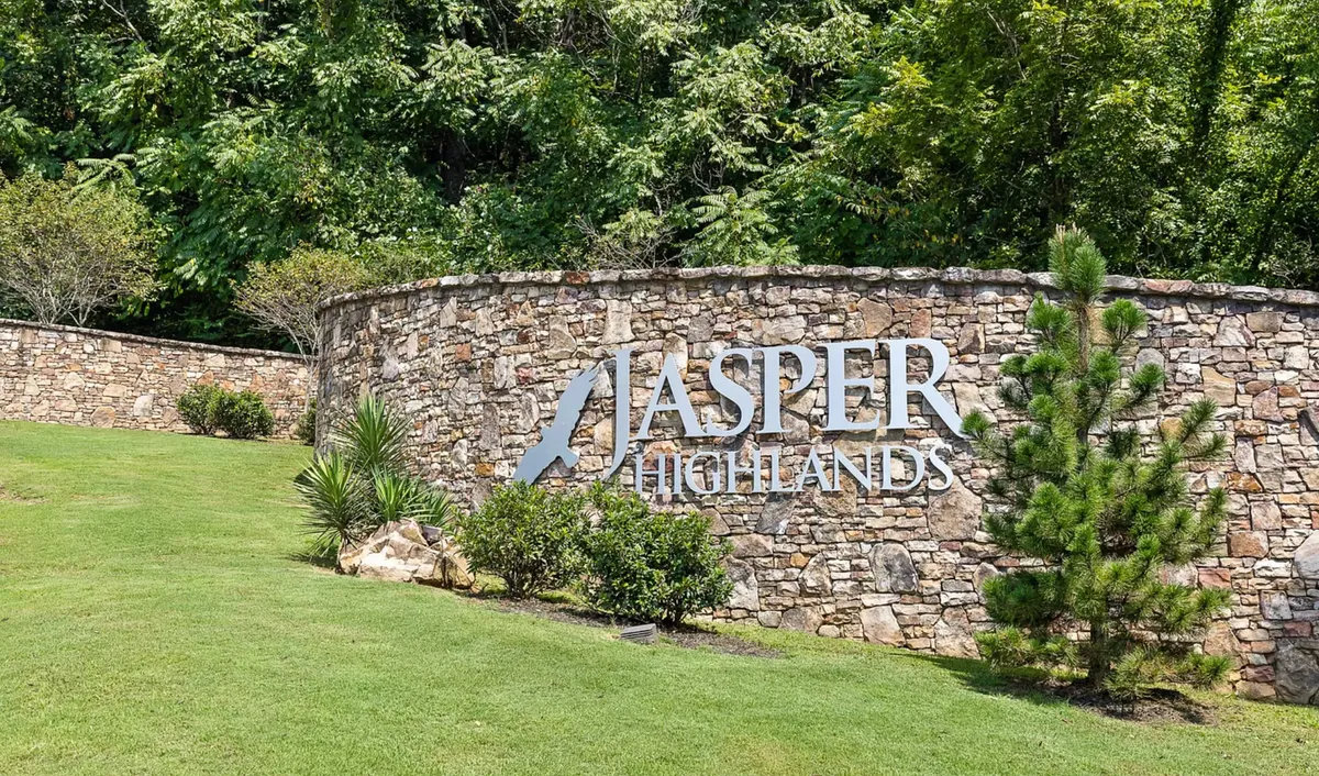 An inviting exterior view of the Jasper Highlands entrance, featuring a stone wall with the community name sign and lush greenery. The image conveys a sense of arrival, welcoming guests into a serene natural setting ideal for exploration and romantic getaways.