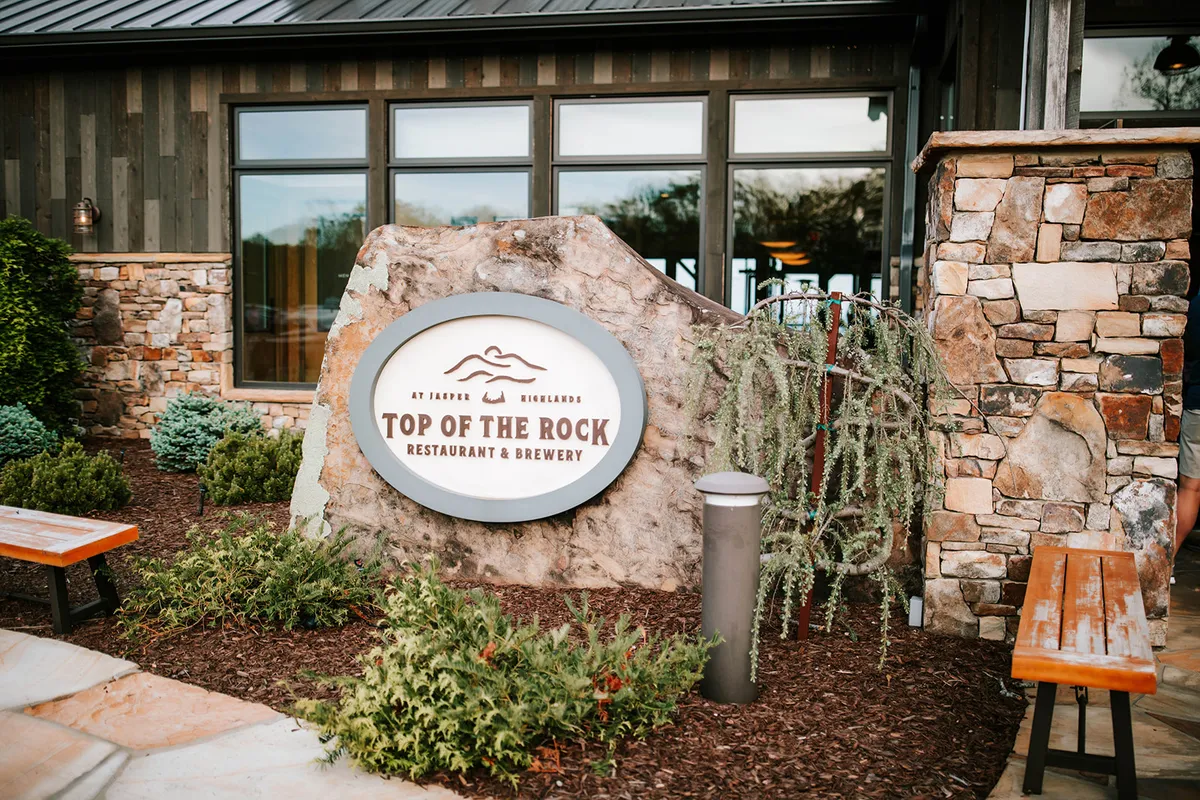The exterior of the Top of the Rock Brewery is featured, with its welcoming restaurant sign, stone walls, benches, and attractive landscaping, all set against a backdrop that hints at social gatherings and outdoor dining.