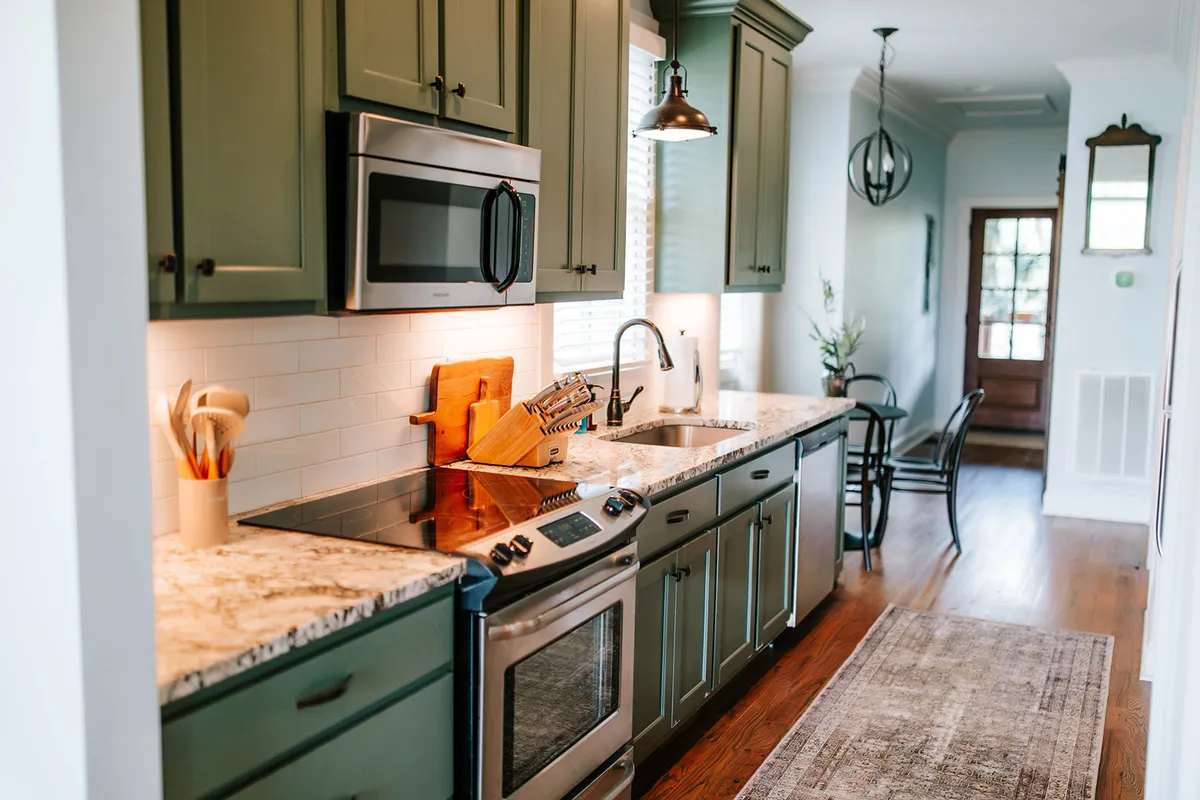 A spacious and modern kitchen featuring a granite countertop, stainless steel appliances, and green cabinetry. A wooden cutting board is placed on the counter, hinting at meal preparation. The warm lighting and thoughtful layout make the space feel inviting for both casual and formal cooking experiences.