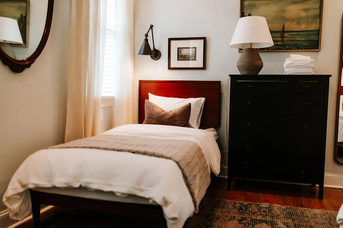 A calm and restful bedroom with a single bed dressed in a clean white duvet, a black dresser, a decorative-framed mirror, and wall art of a tranquil seascape. Natural light gently illuminates the space, underscoring its serene atmosphere.